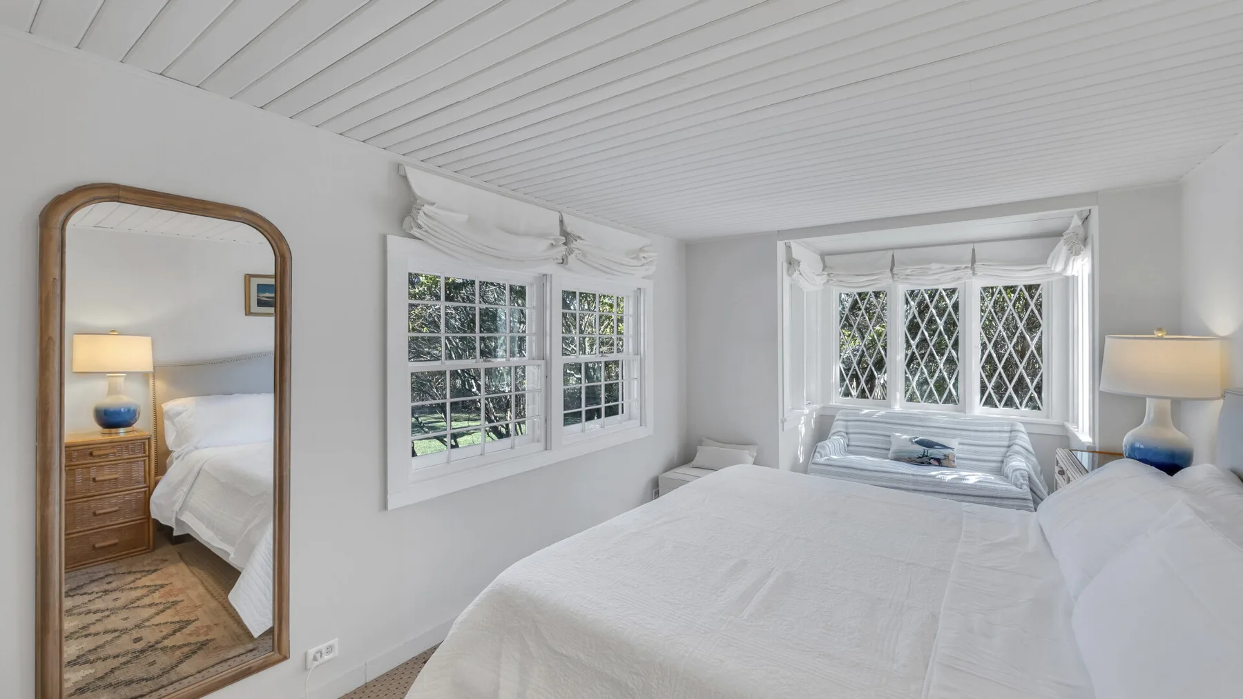 A bright bedroom with white bedding, multiple windows, and a wood-framed mirror.