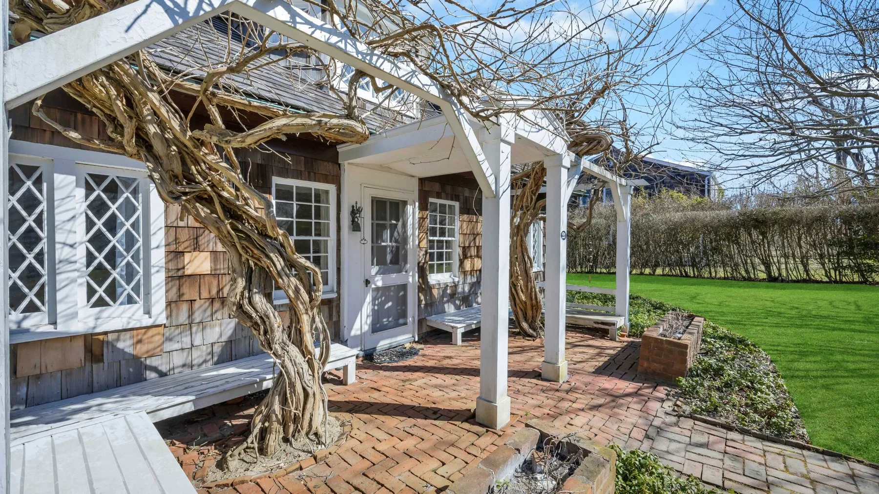A brick patio beside a shingled house sits beneath a white pergola.