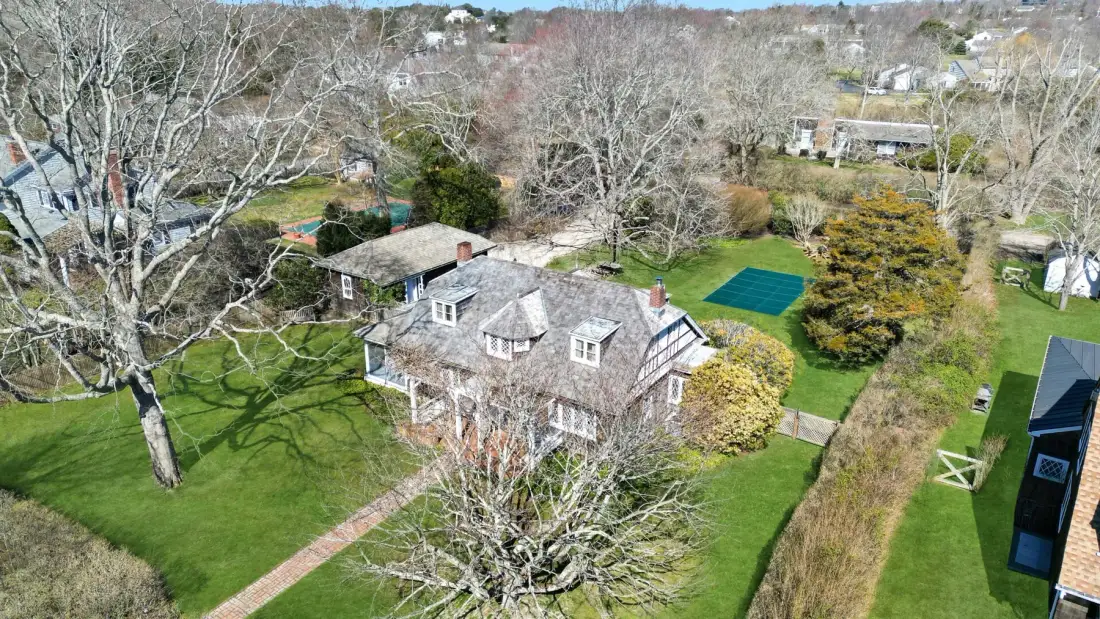 An aerial view shows a house surrounded by lawns, trees, and neighboring homes.
