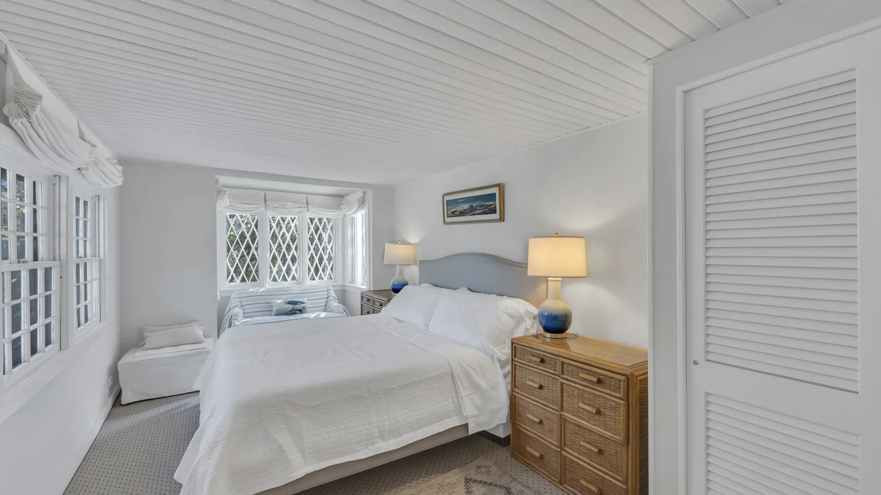 A bright bedroom with white bedding and a wooden dresser beside the bed.