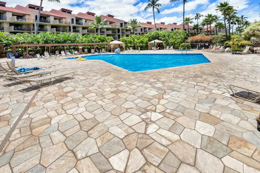A swimming pool and paved deck are surrounded by lounge chairs and palm trees.