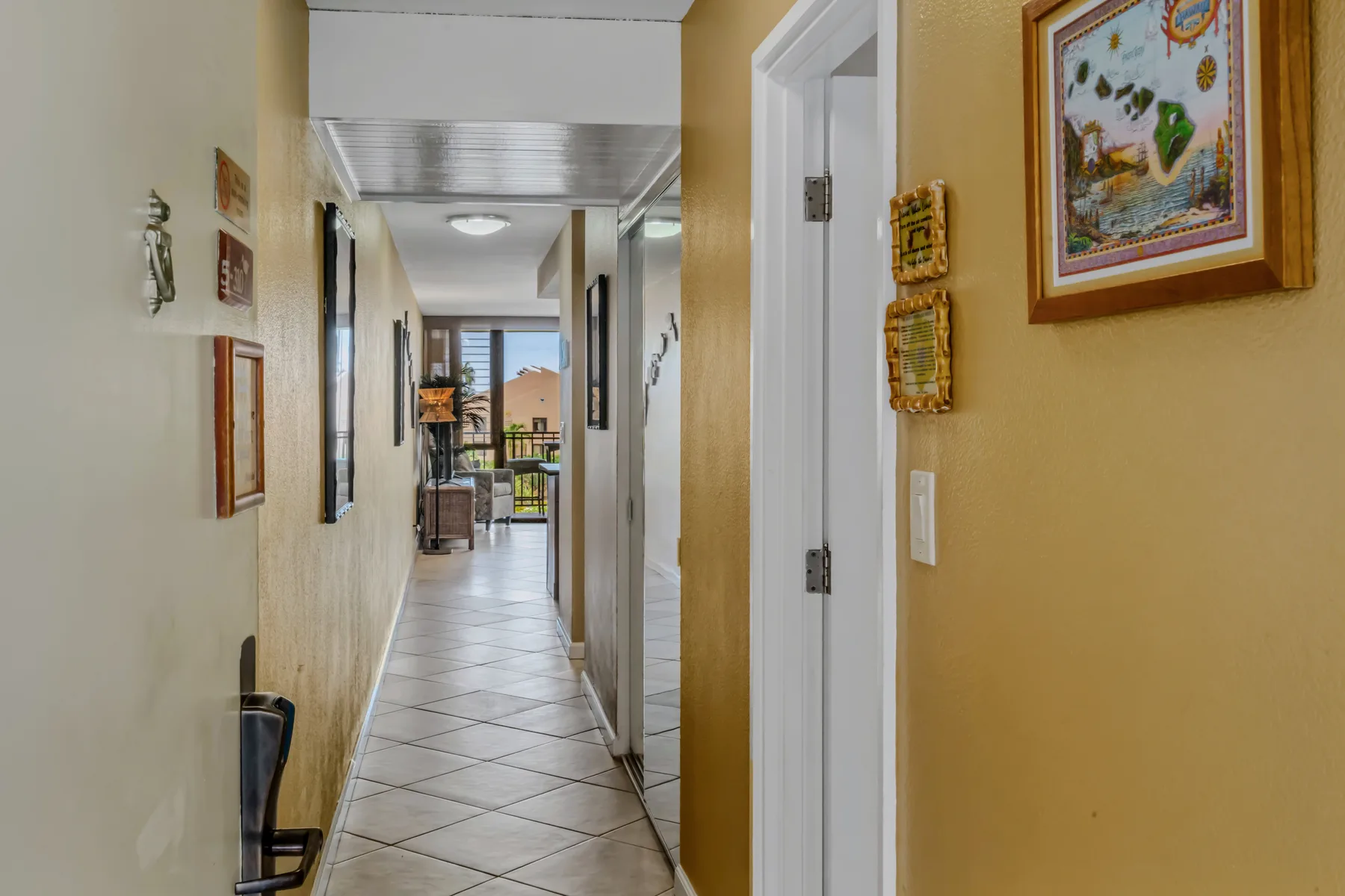 A tiled hallway with framed artwork and doorways leading toward a bright room.