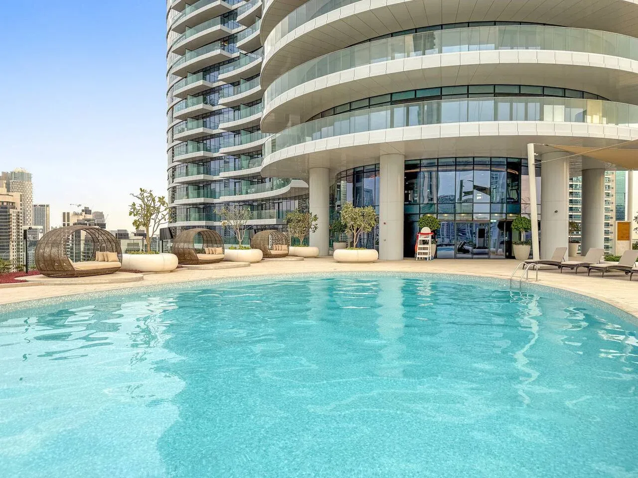 A rooftop swimming pool sits beside a curved modern building and outdoor seating.