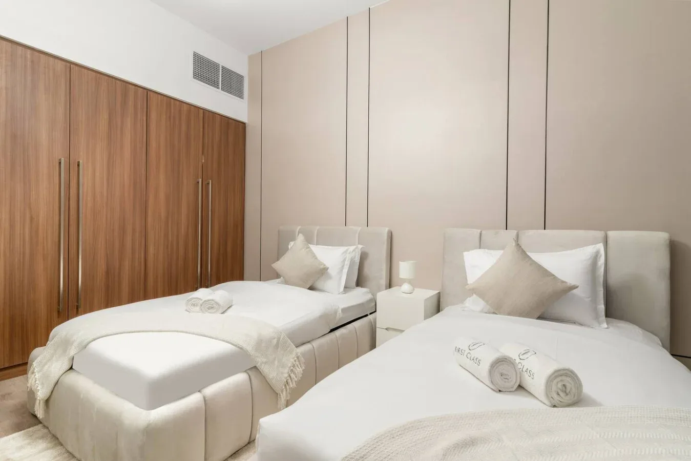 A bedroom with two beds, neutral bedding, and built-in wooden wardrobes.