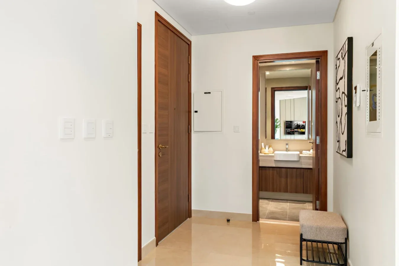 A bright hallway with a wooden door, a bench, and a bathroom visible beyond.