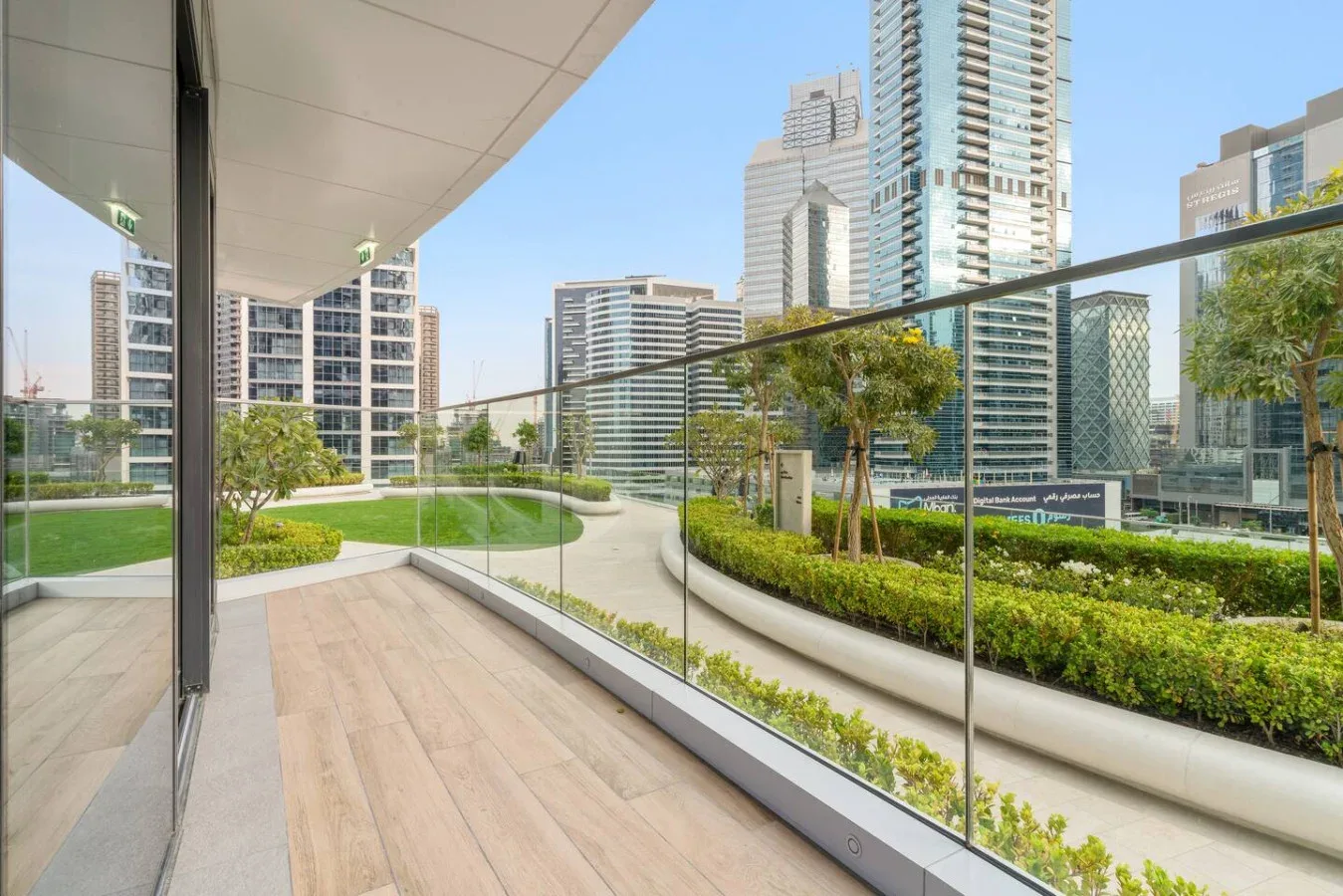 A modern outdoor terrace with glass railings overlooks landscaped greenery and high-rise buildings.
