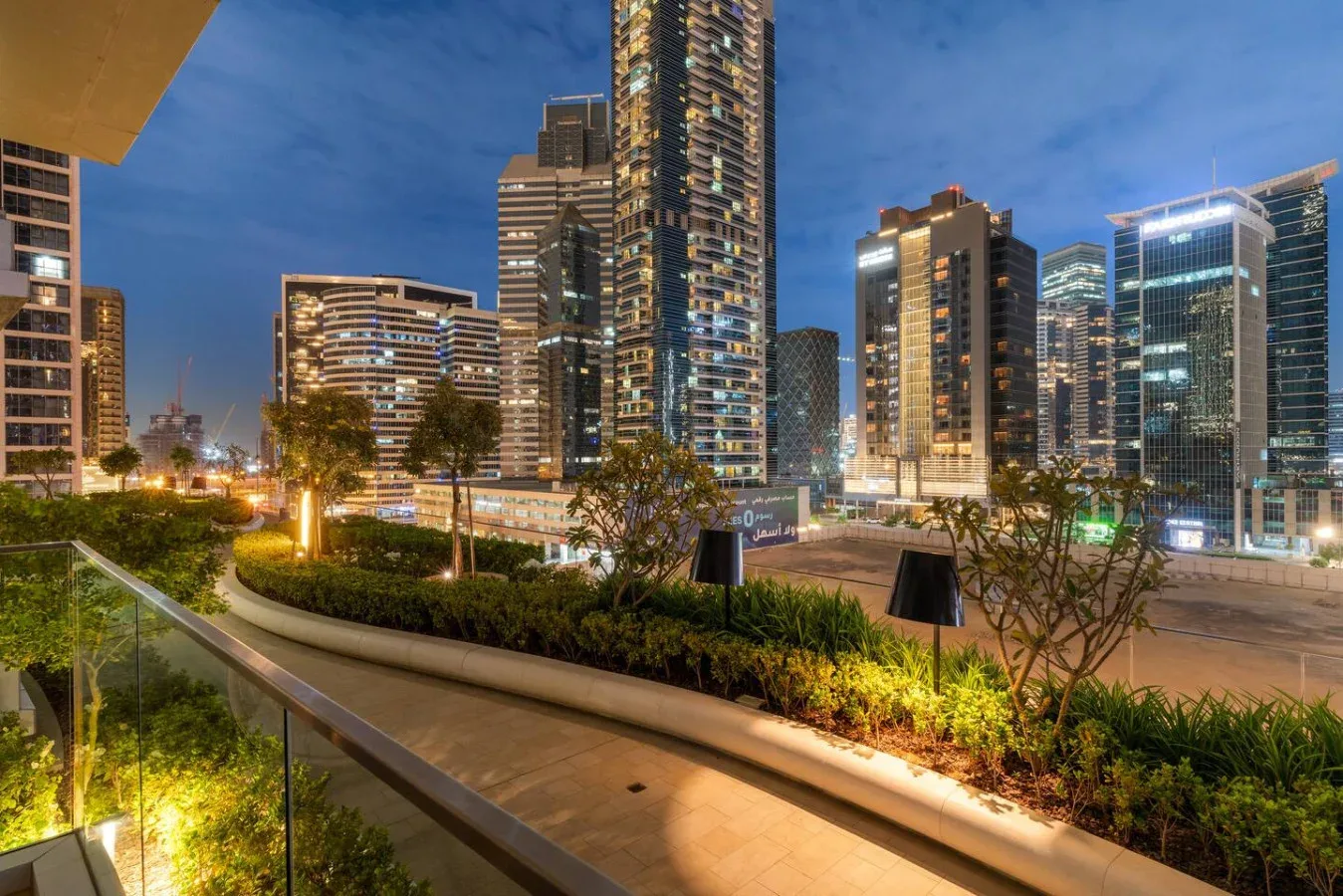 A landscaped pedestrian walkway with planted beds sits in front of illuminated high-rise buildings.