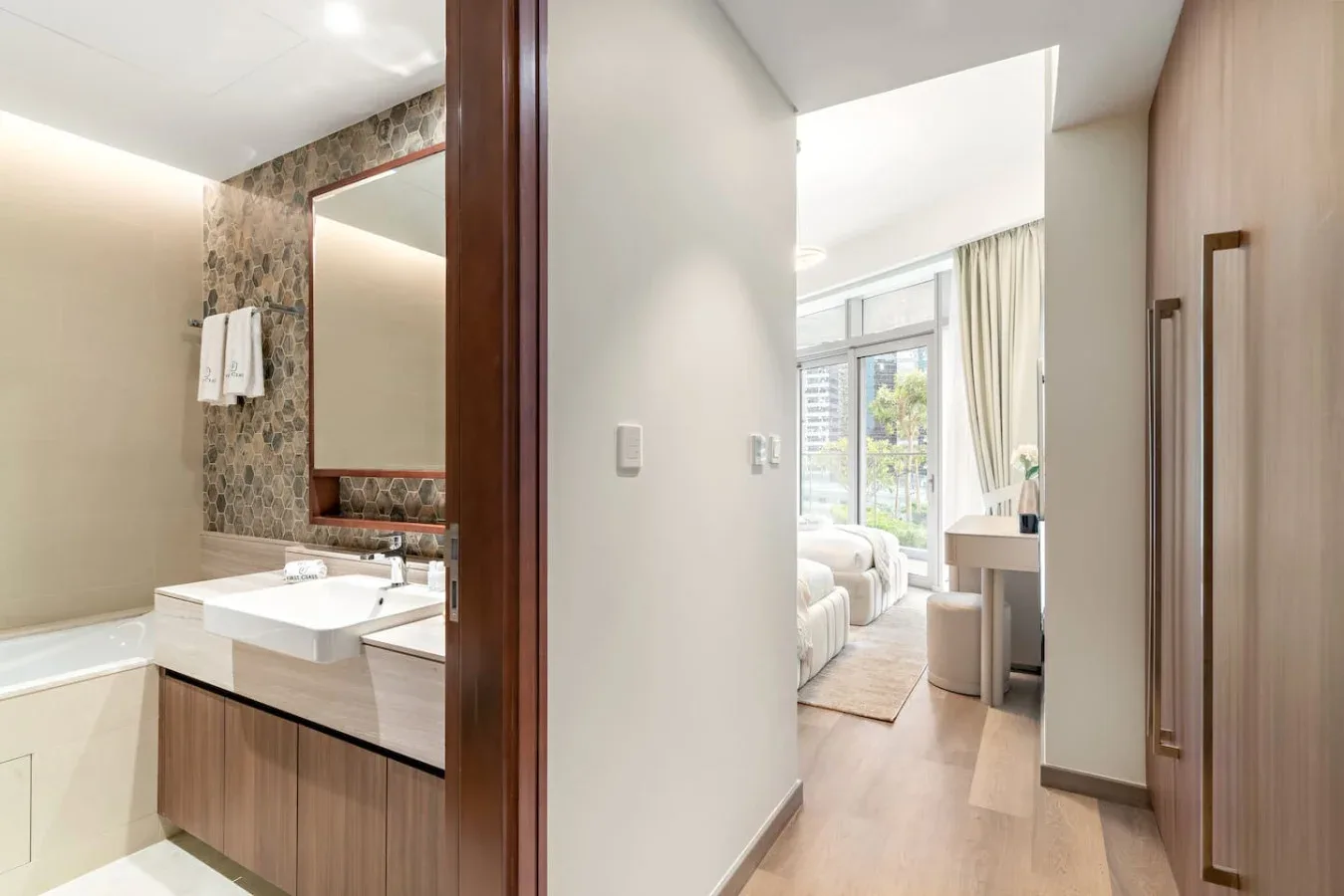 A bathroom opens onto a bright bedroom with a bed and large windows.