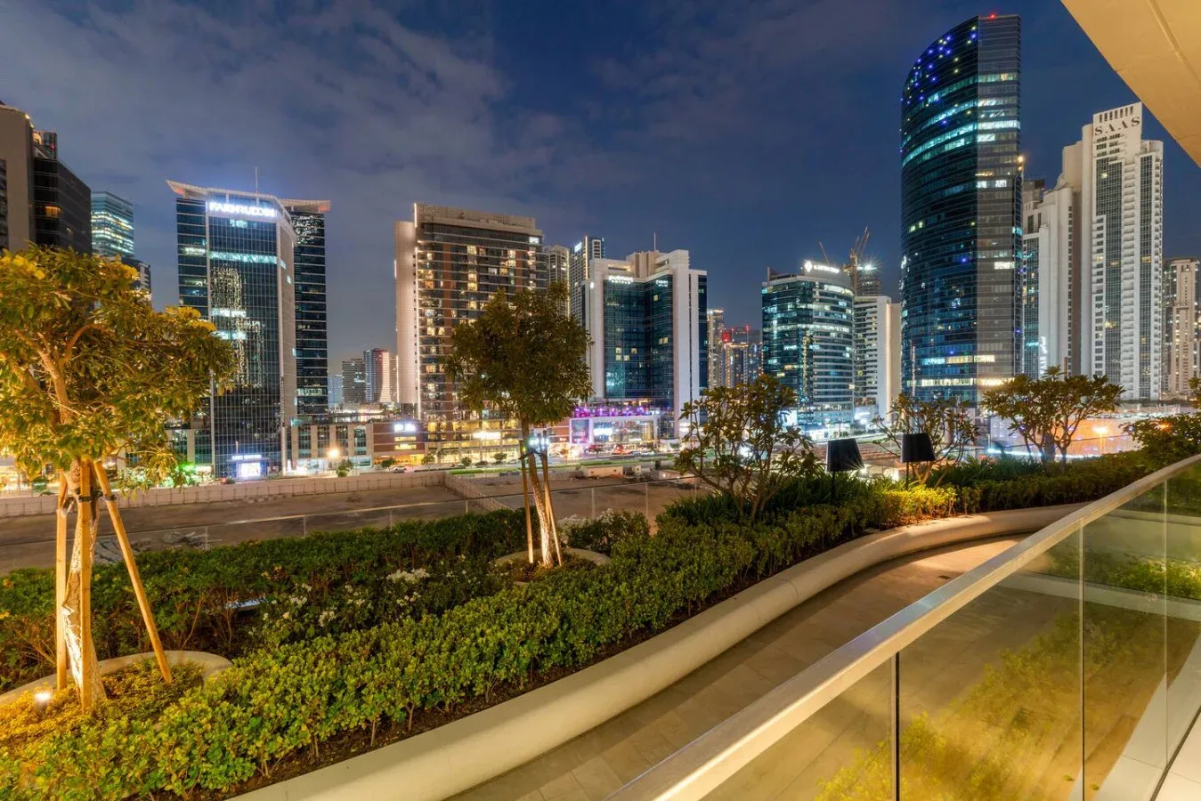 A landscaped pedestrian path overlooks illuminated city skyscrapers at night.