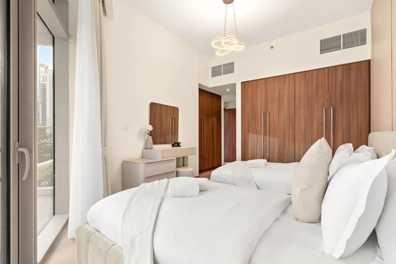 A bright bedroom with white bedding, wooden wardrobes, and a large window.