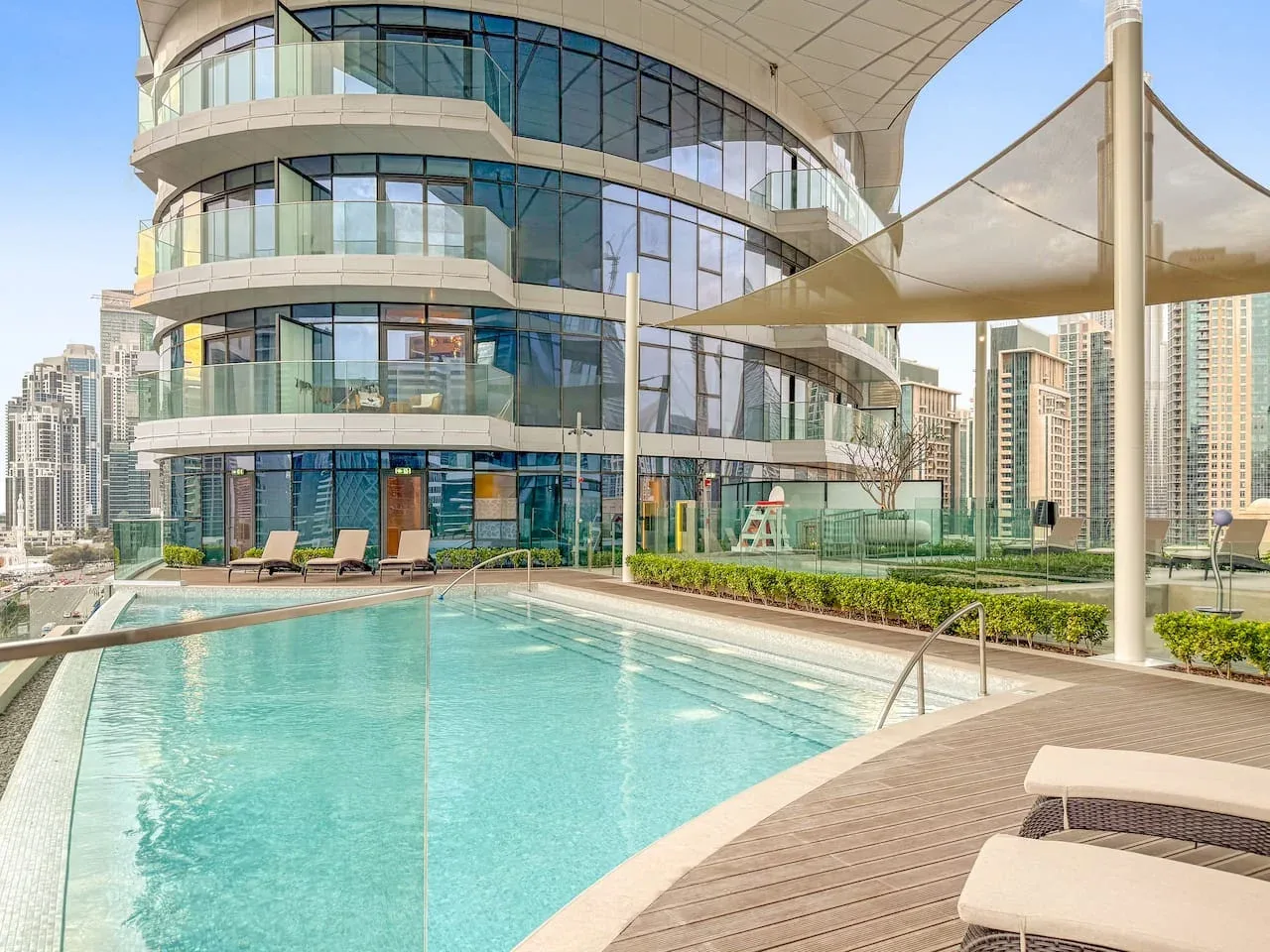 A rooftop swimming pool beside a curved glass building and shaded seating area.