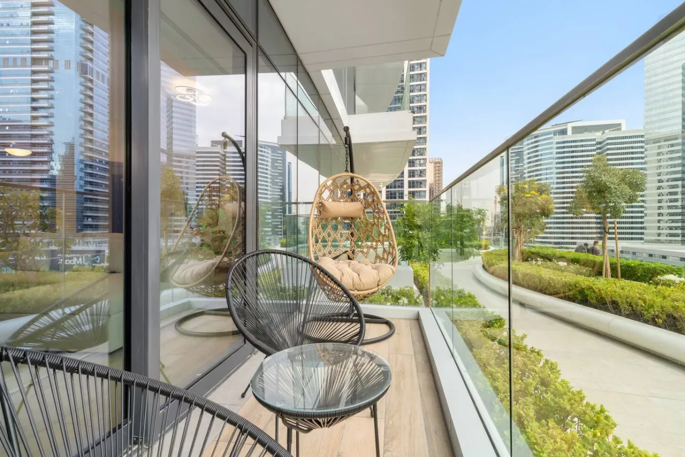 A glass-sided balcony has seating and overlooks landscaped grounds and nearby buildings.