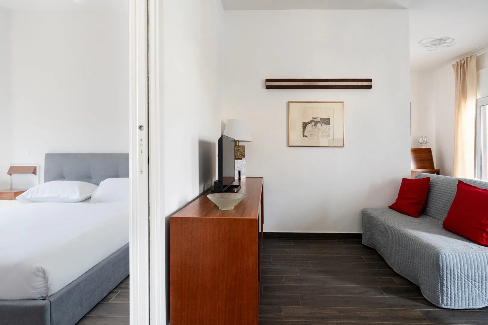Un dormitorio luminoso con zona de estar, cama, televisor y sofá gris.