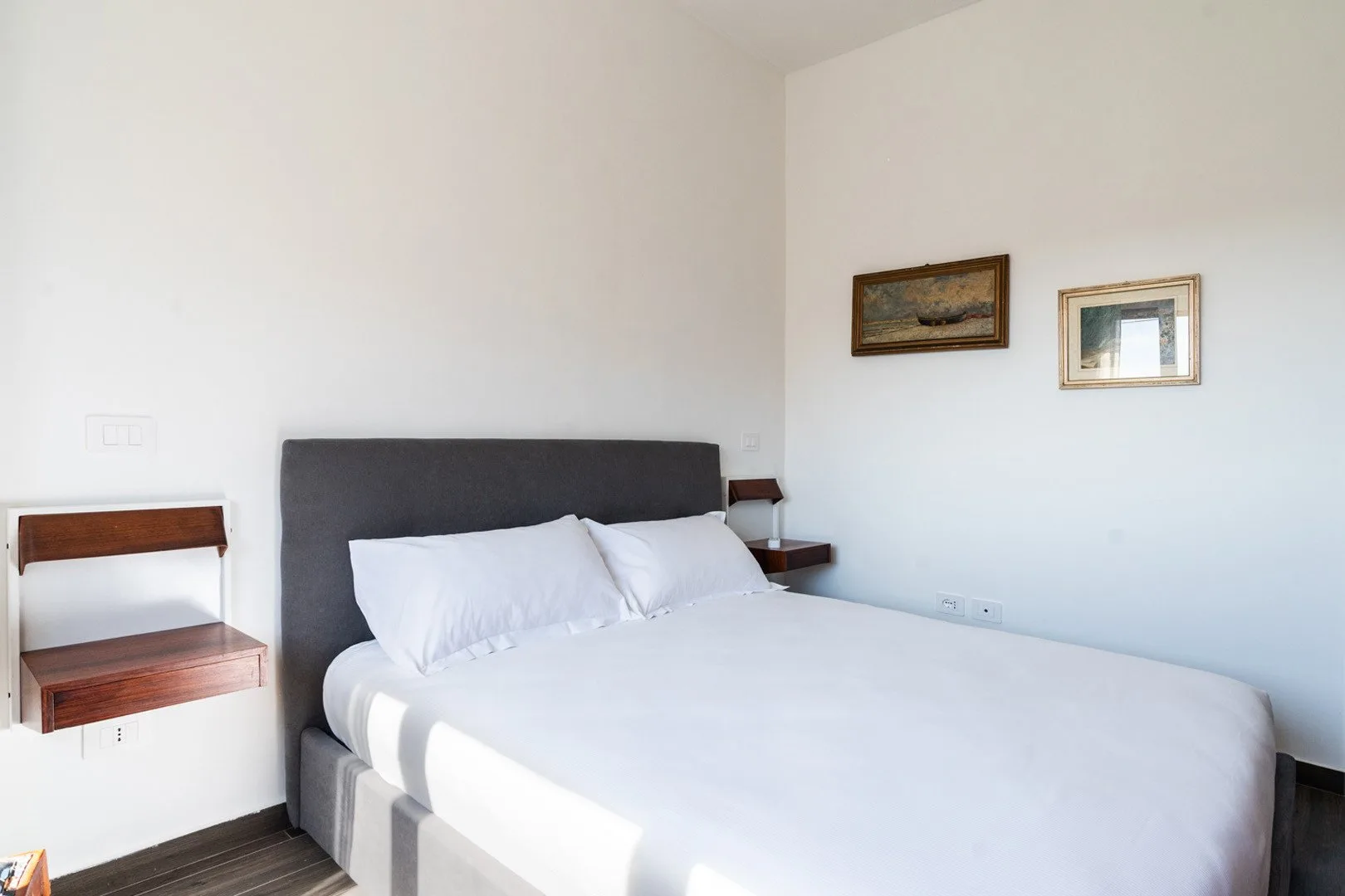 Un dormitorio luminoso con una cama grande, ropa de cama blanca y cuadros en la pared.