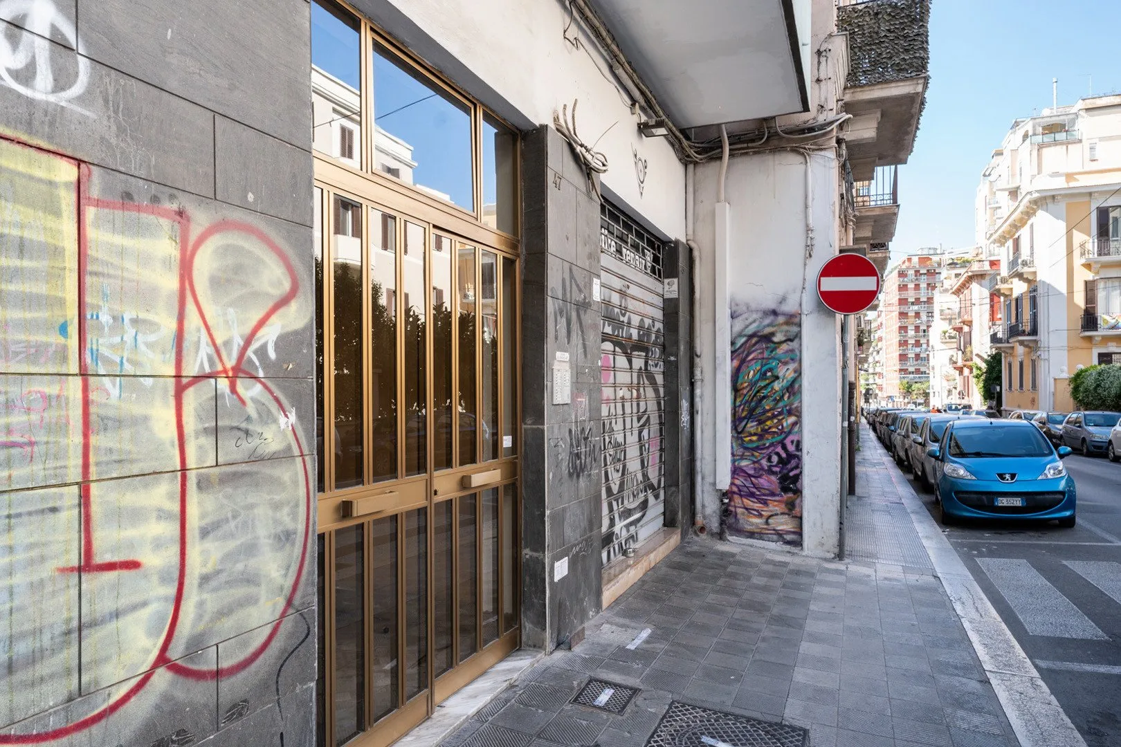 Una acera urbana pasa junto a escaparates con grafitis y coches estacionados.