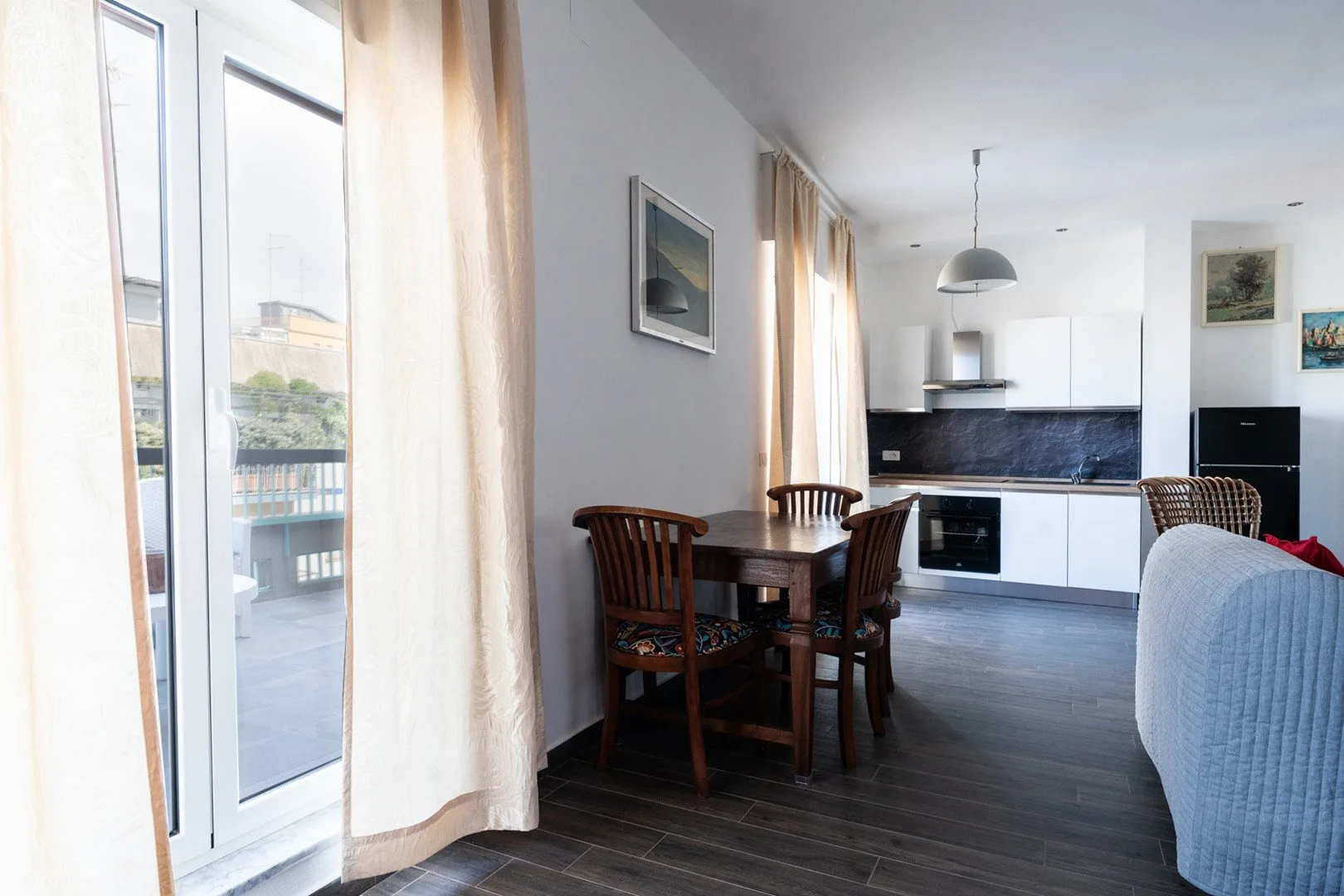 Una luminosa zona de comedor y cocina con una mesa de madera junto a ventanas altas.