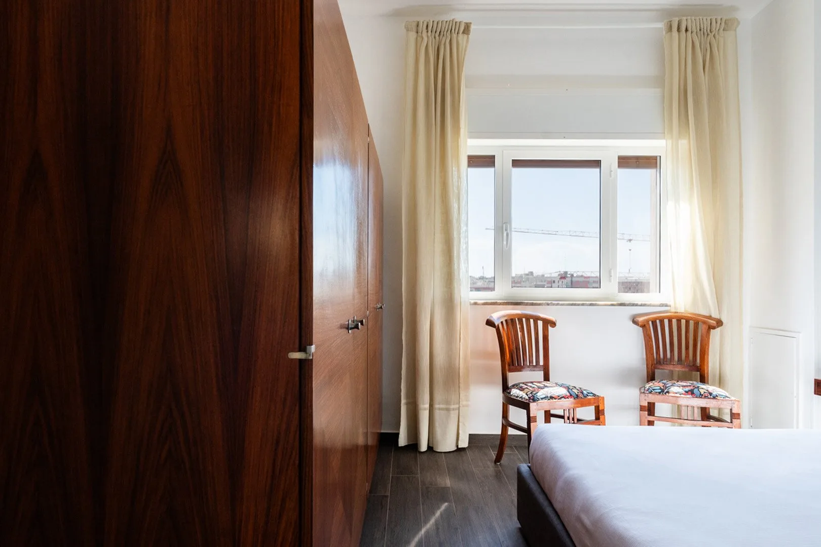 Un dormitorio con una ventana grande, cortinas claras y dos sillas de madera.