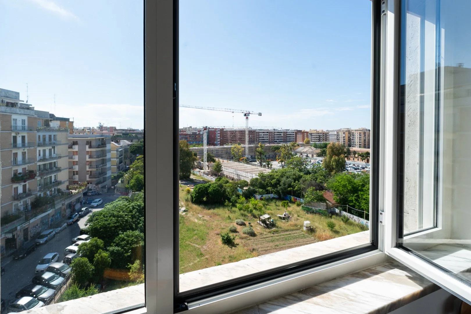 Una ventana abierta da a edificios de apartamentos, árboles y una obra en construcción.