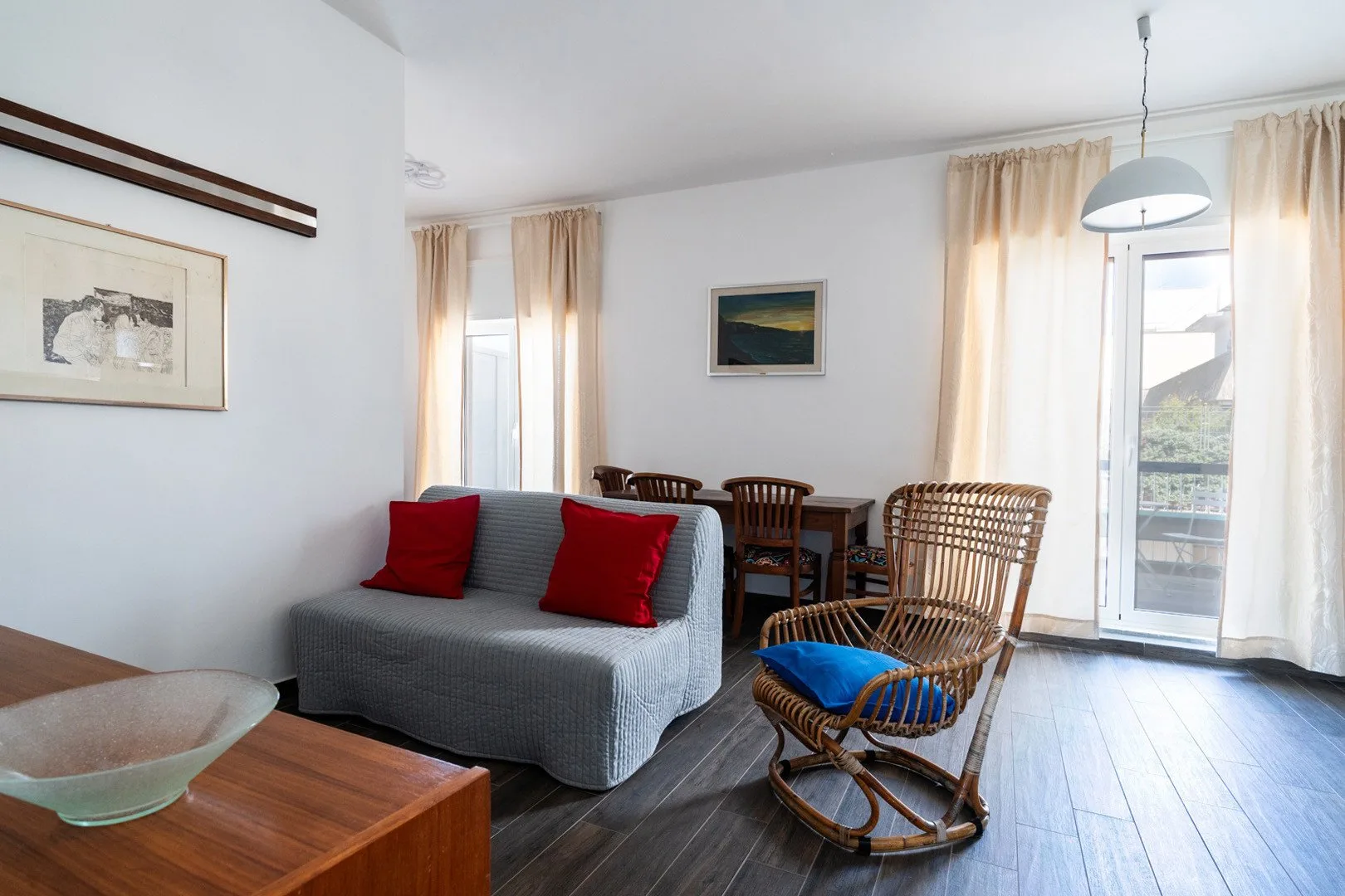 Una sala de estar con un sofá gris y asientos de mimbre junto a ventanas altas.