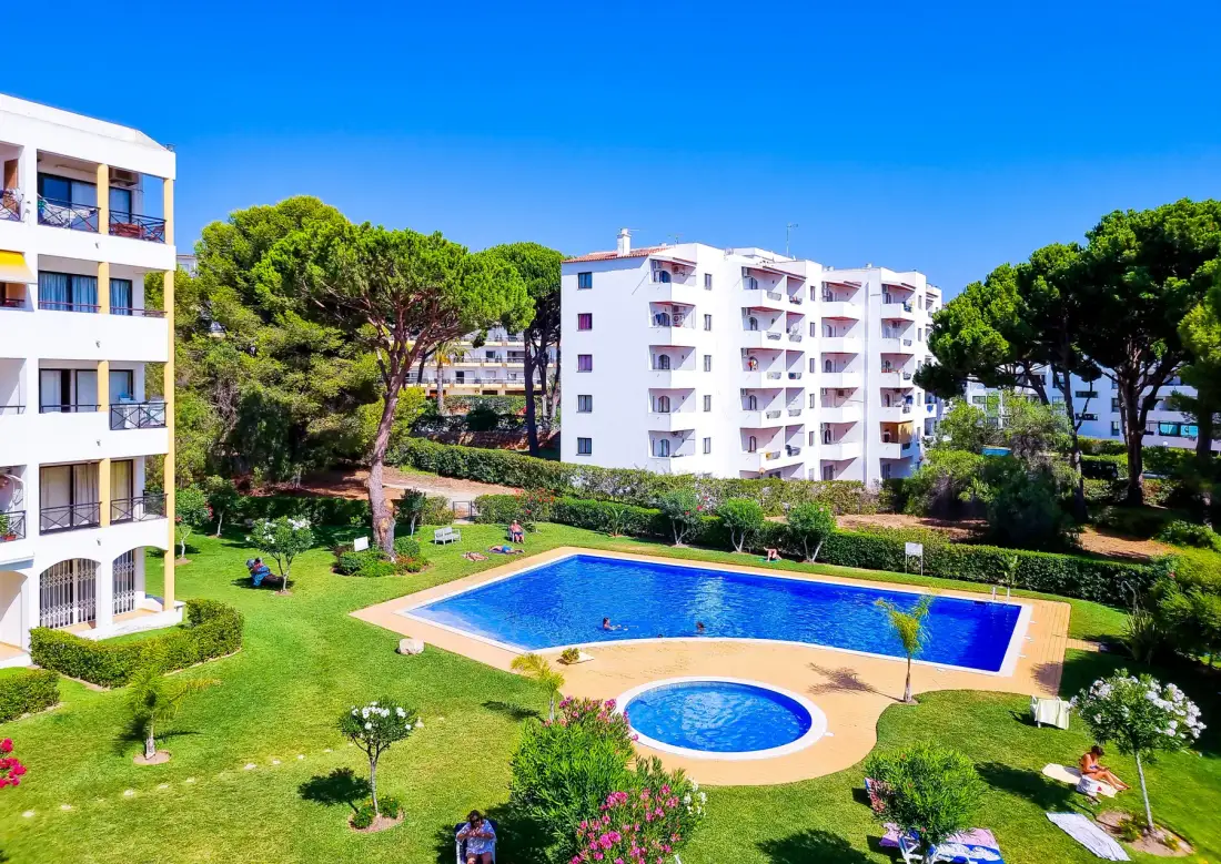 A landscaped courtyard with a swimming pool sits among white apartment buildings and trees.