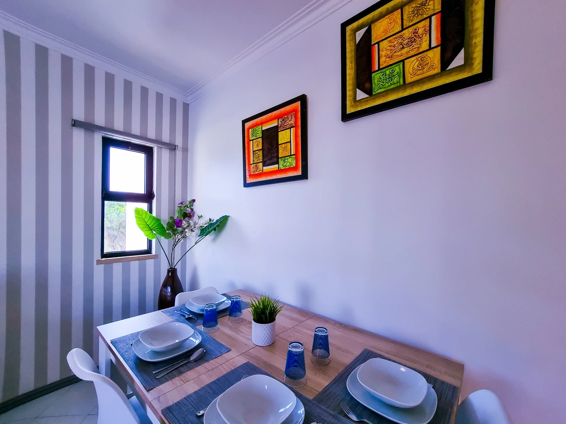 A dining area with a set table, colorful framed art, and a striped wall.