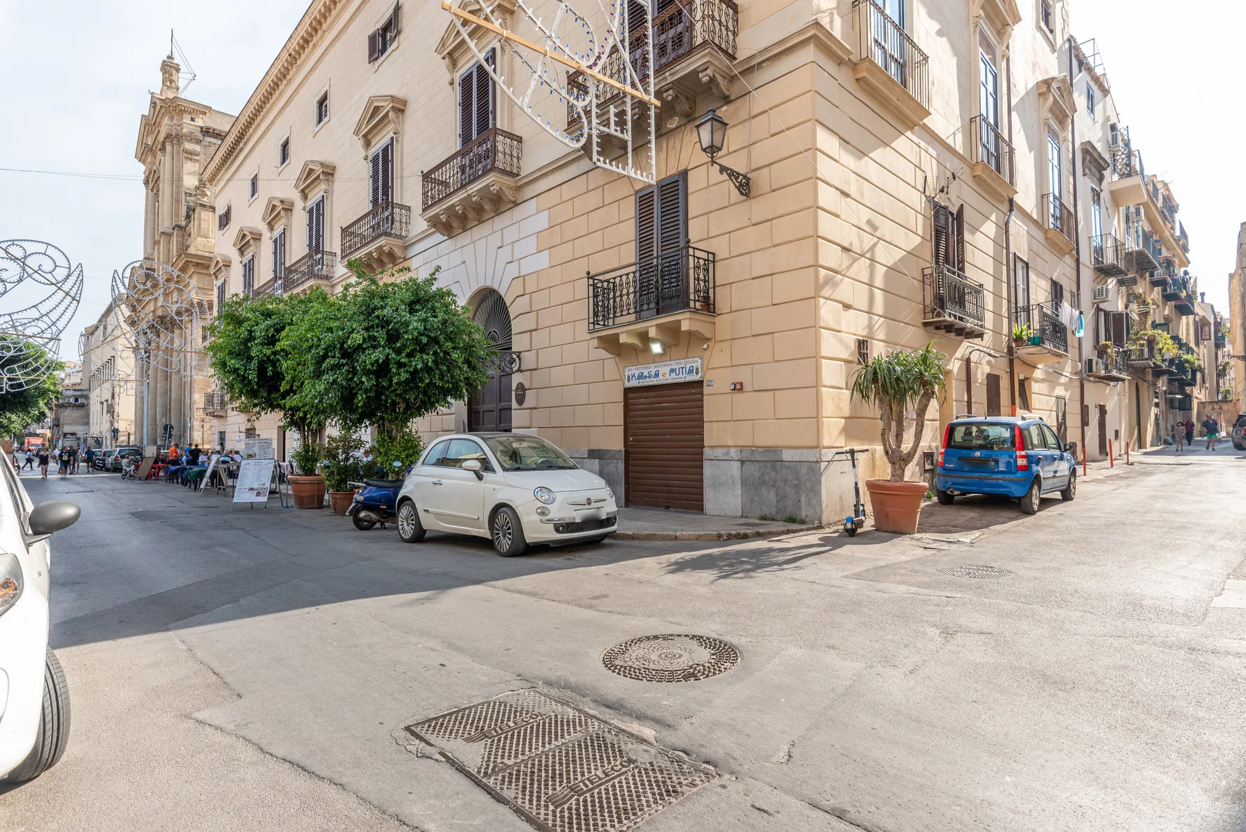 Una esquina soleada con edificios claros, coches estacionados y plantas en macetas.