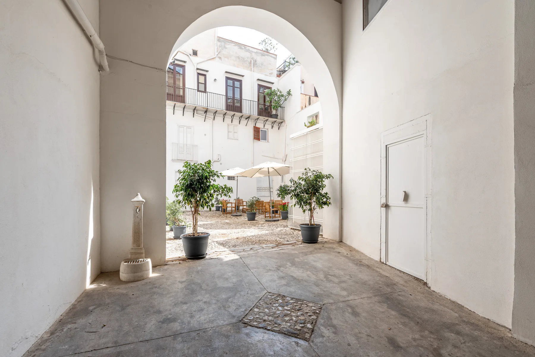 Un patio luminoso queda enmarcado por un gran arco, con árboles en macetas y asientos al aire libre.