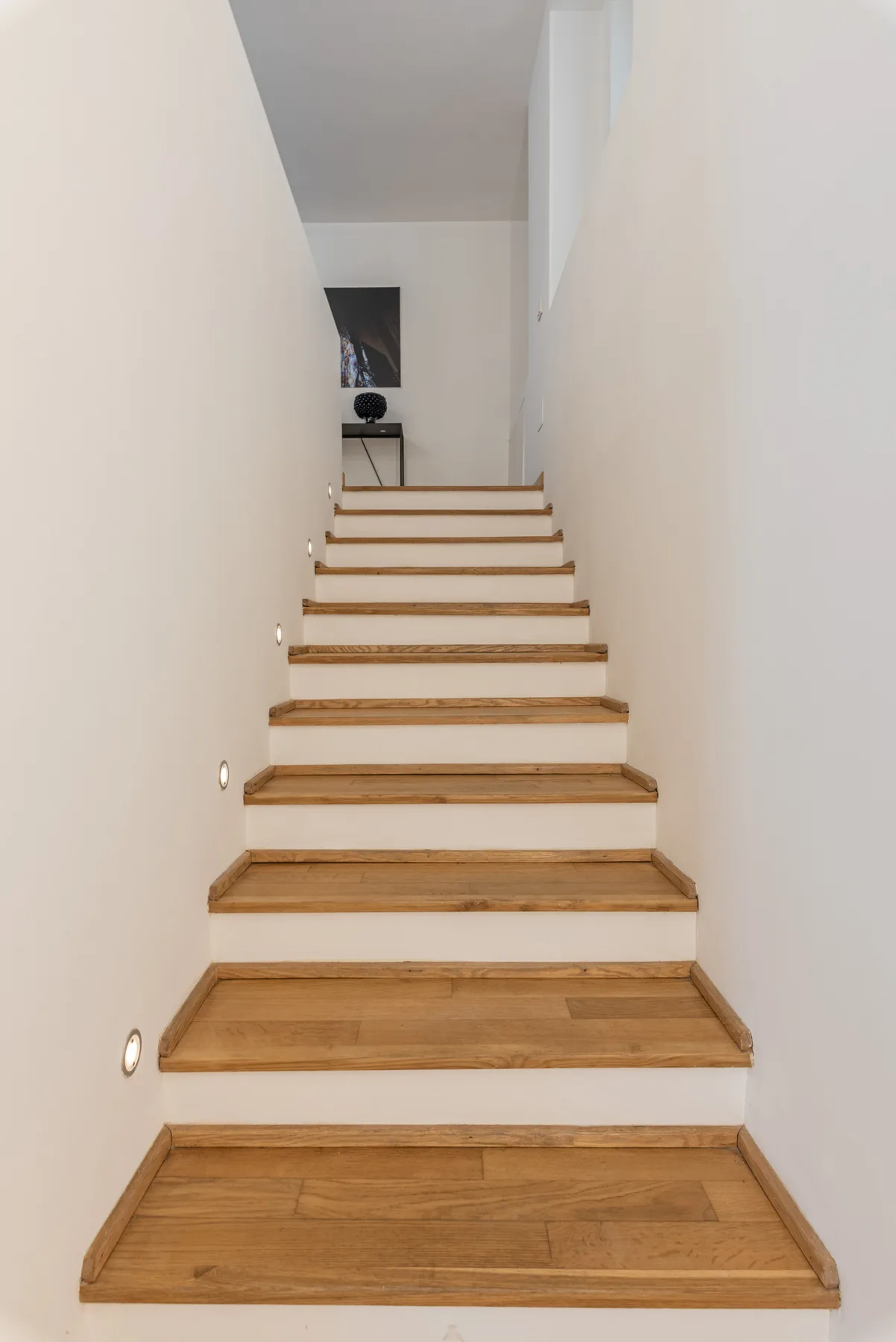 Una escalera interior con peldaños de madera sube entre paredes blancas.