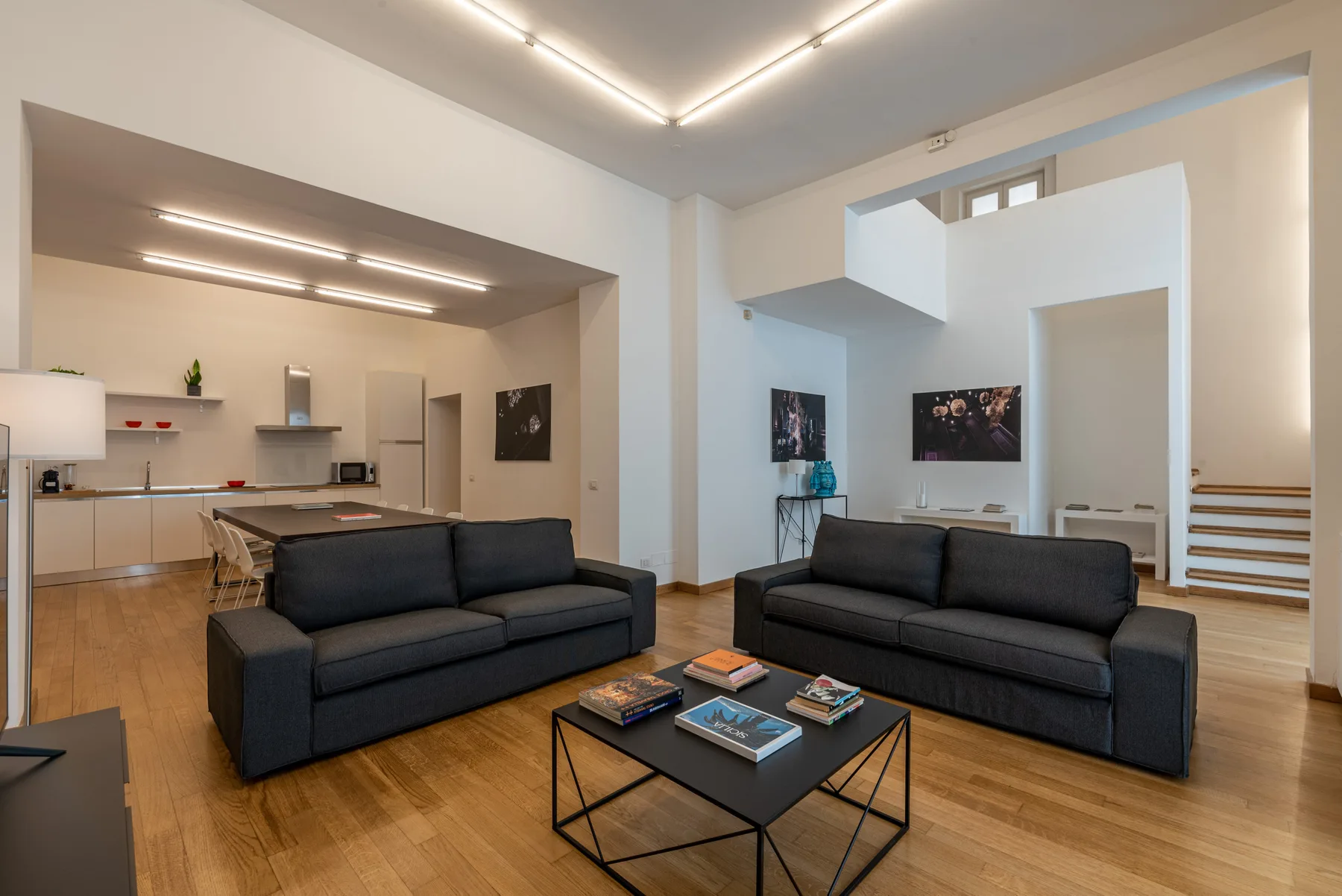 Una luminosa sala de estar abierta con dos sofás oscuros y una cocina al fondo.
