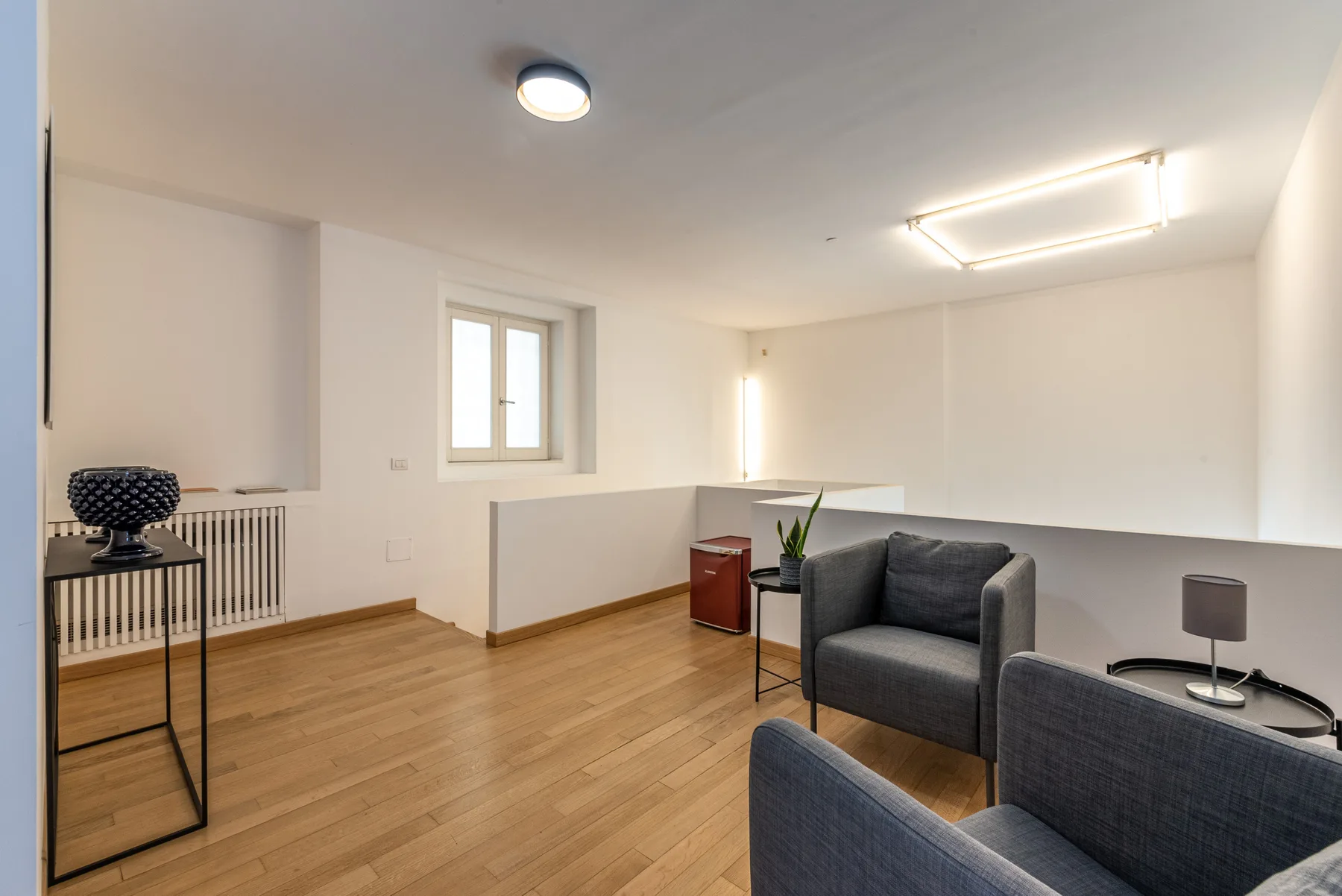 Una luminosa zona de estar con asientos grises, una ventana y suelo de madera clara.