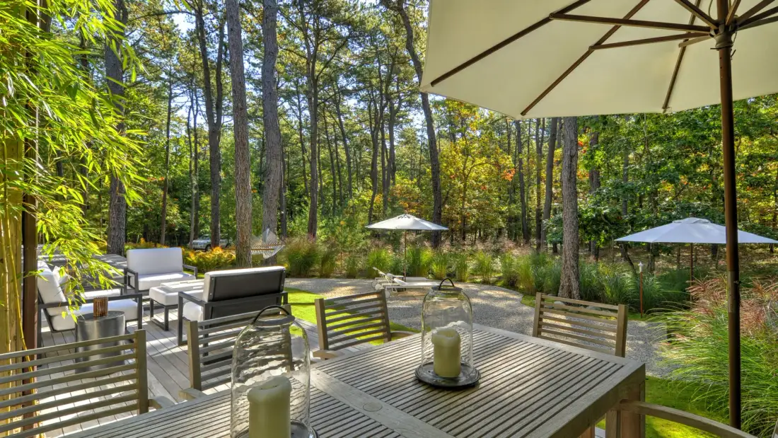 An outdoor patio with a slatted table and seating overlooks a wooded garden.
