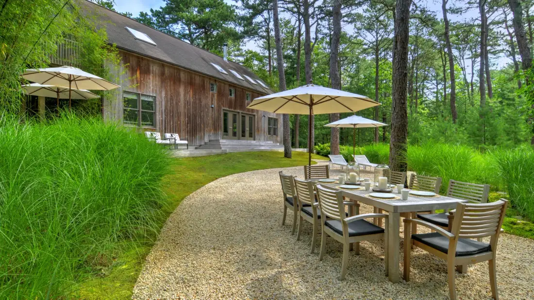 A gravel patio with outdoor dining tables and umbrellas sits beside a wood-sided building.