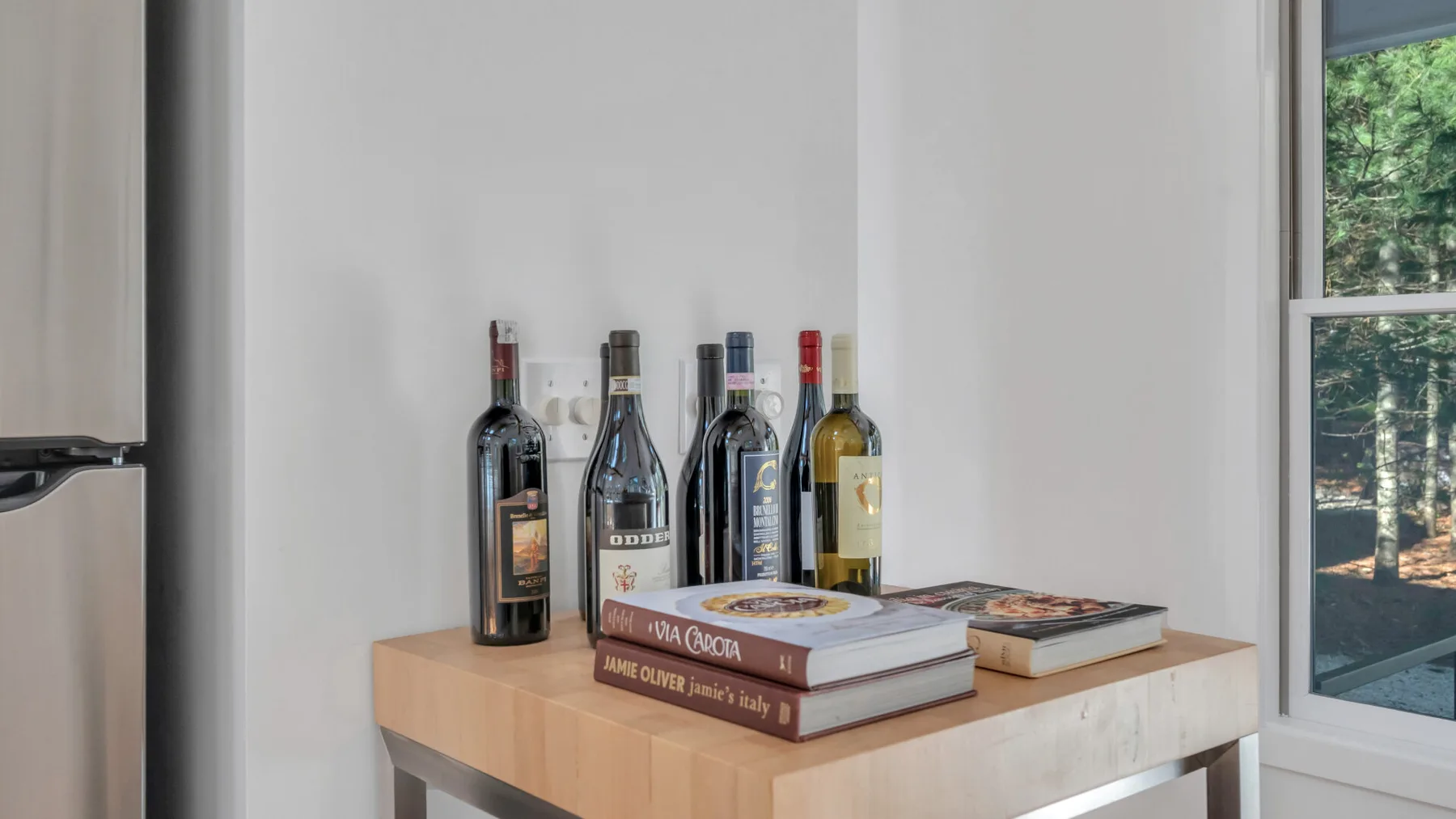 A wooden table displays several wine bottles and books beside a window.