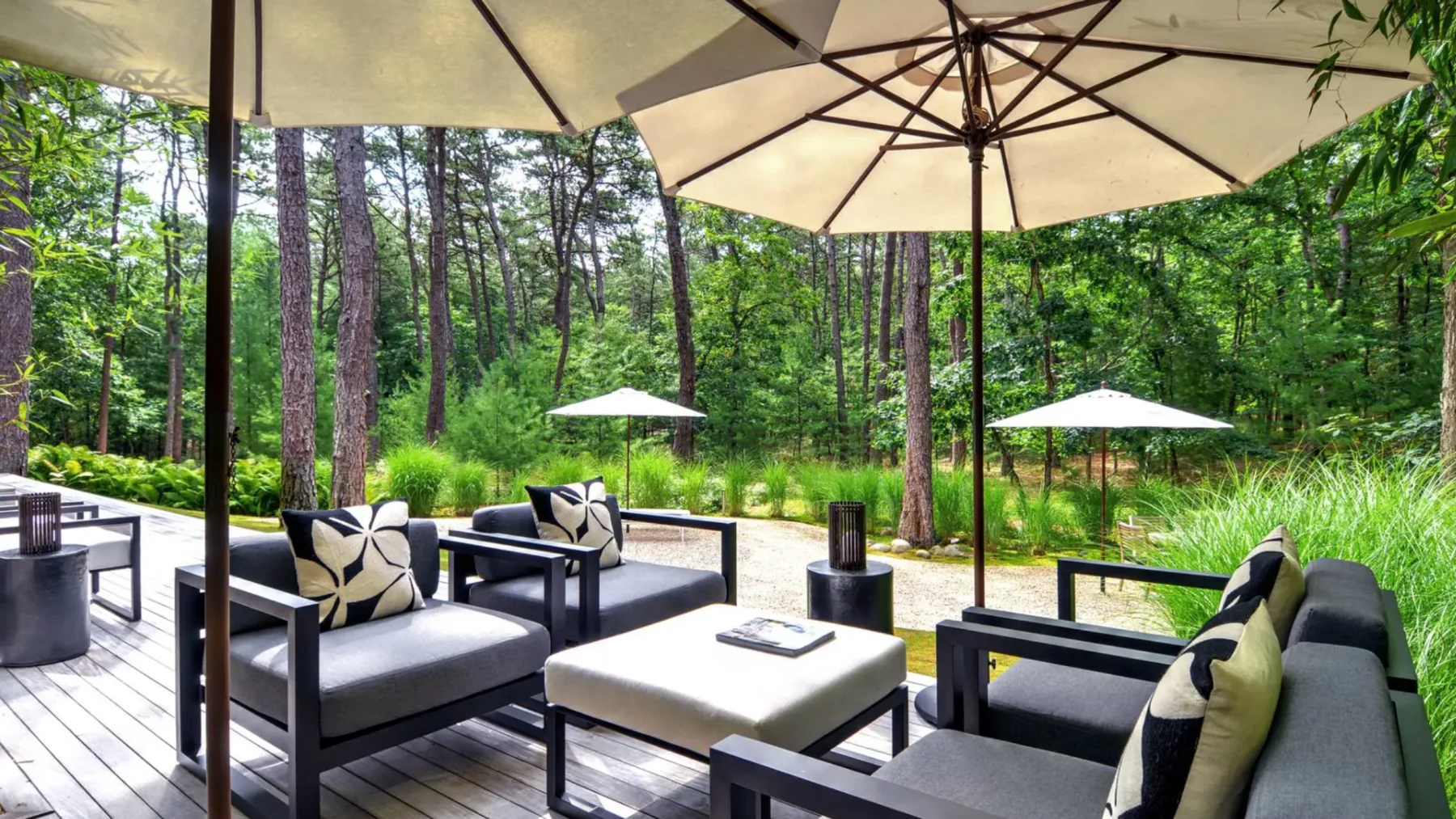 An outdoor seating area with cushioned furniture and large umbrellas beside a wooded setting.
