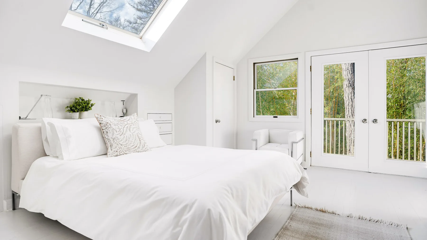 A bright bedroom with a white bed, skylight, and glass doors opening onto greenery.