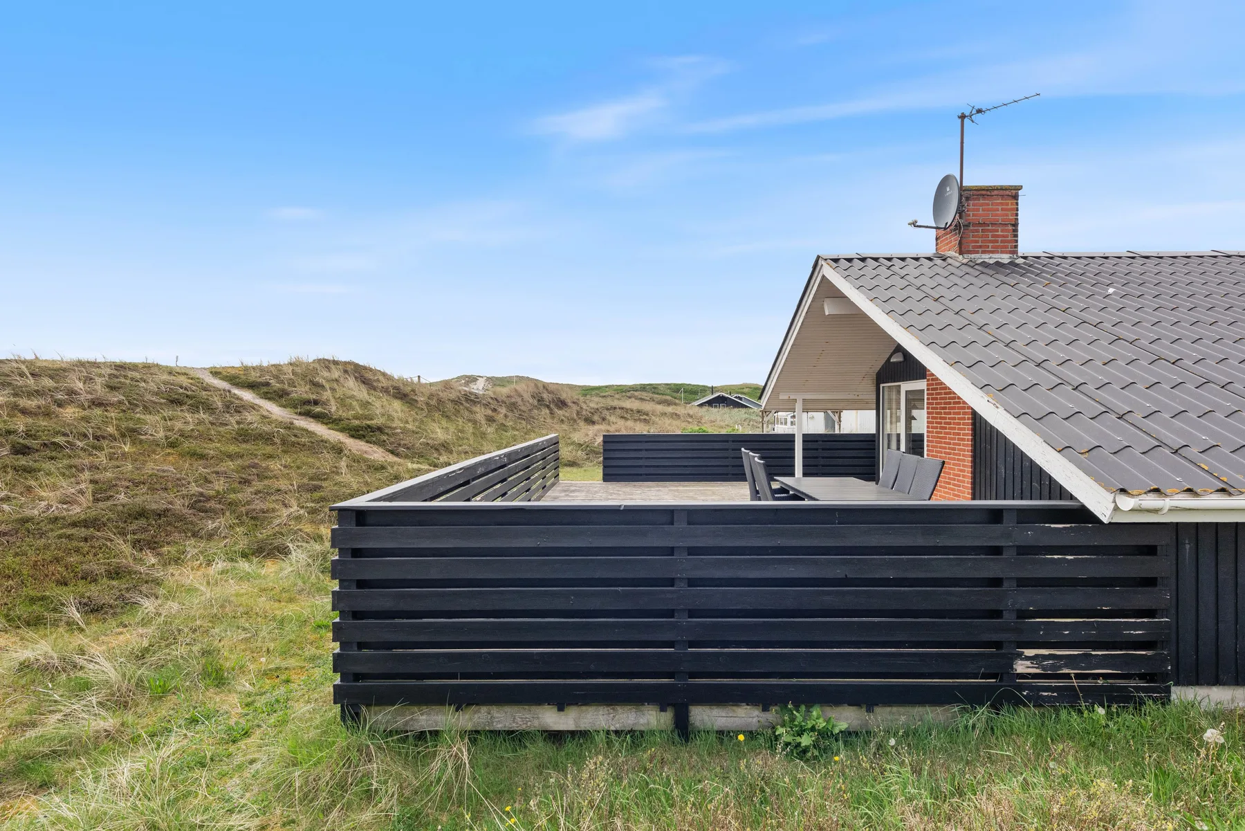 Una terraza exterior cercada está junto a una casa, con dunas cubiertas de hierba al fondo.