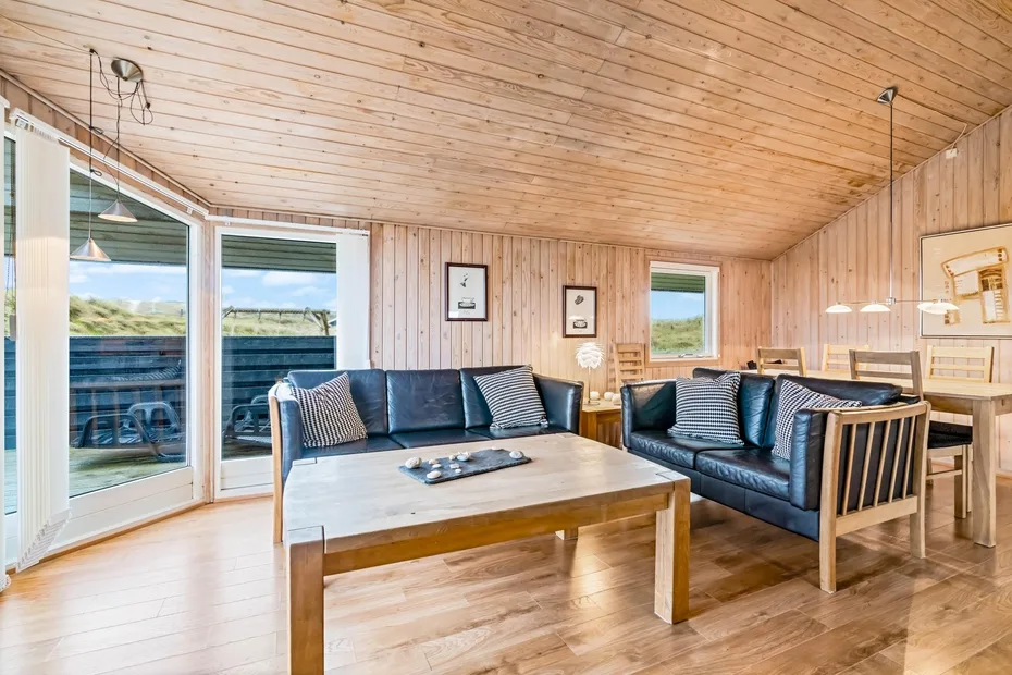 Sala con paredes de madera, asientos oscuros y una mesa de centro junto a grandes ventanas.