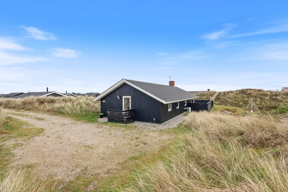 Una casa oscura se encuentra entre dunas cubiertas de hierba, junto a un camino de arena.