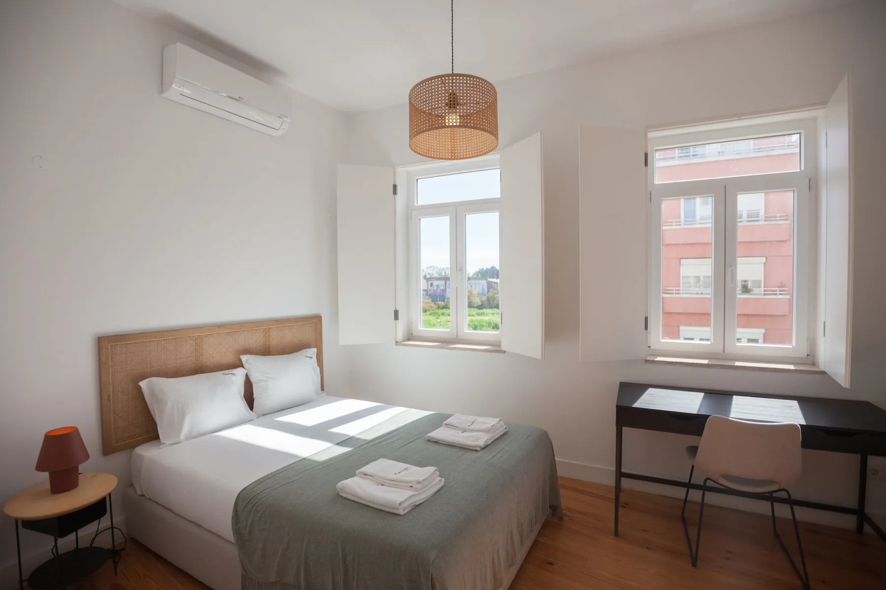 A bedroom with a neatly made bed, bedside table, and desk by the windows.