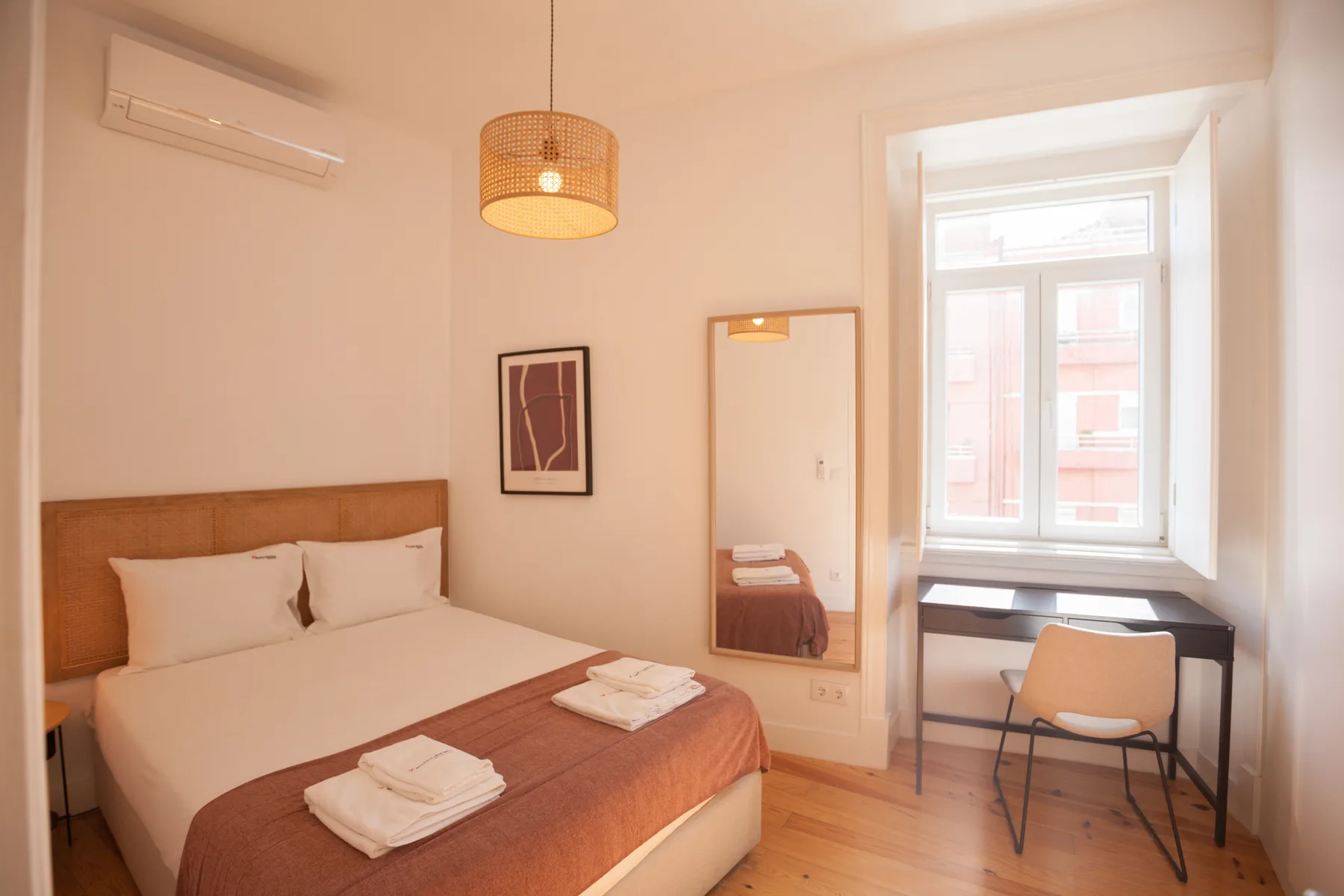 A bedroom with a bed, wall mirror, and desk beside a window.