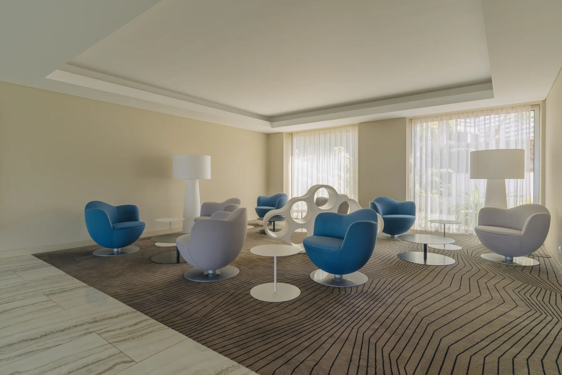 A bright living room with blue seating, a patterned rug, and large curtained windows.
