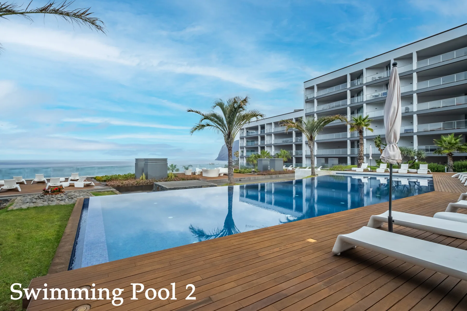 An outdoor swimming pool beside a wooden deck, palm trees, and a multi-story building.