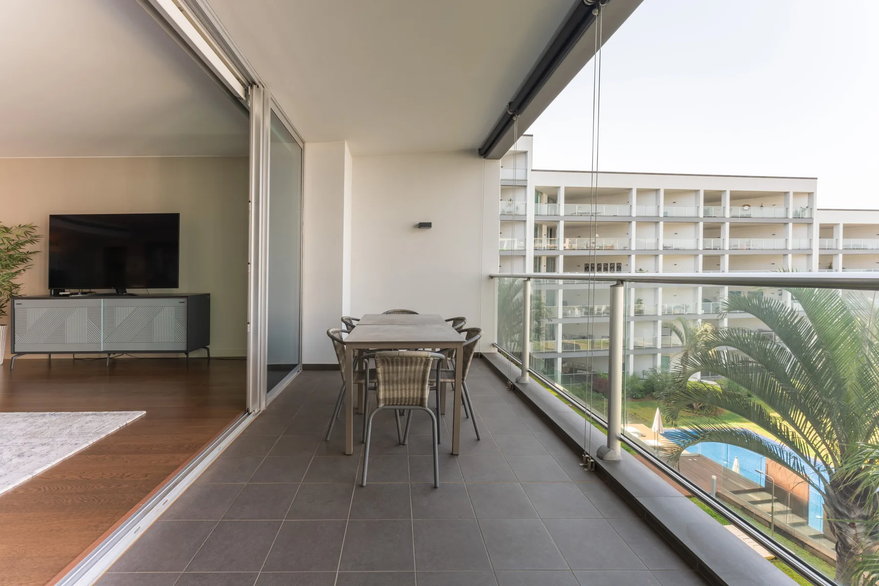 A covered balcony has a dining table beside glass doors overlooking a pool.
