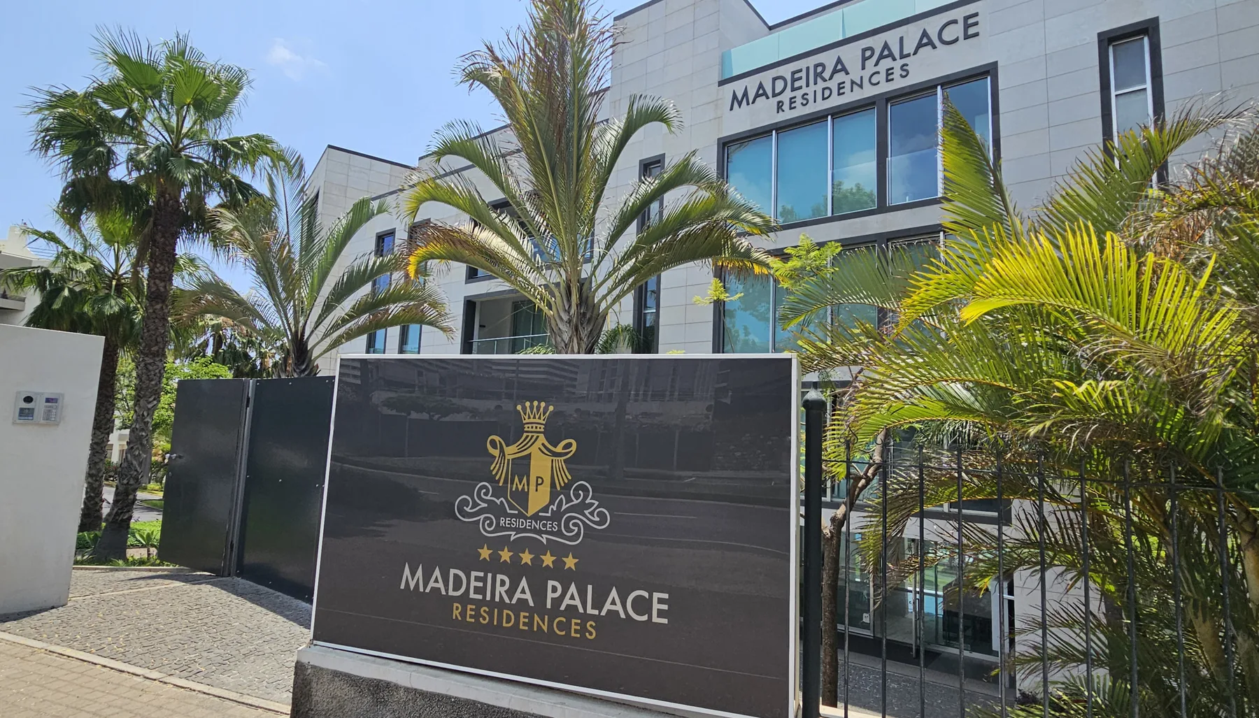 A modern building entrance with palm trees and a sign for Madeira Palace Residences.