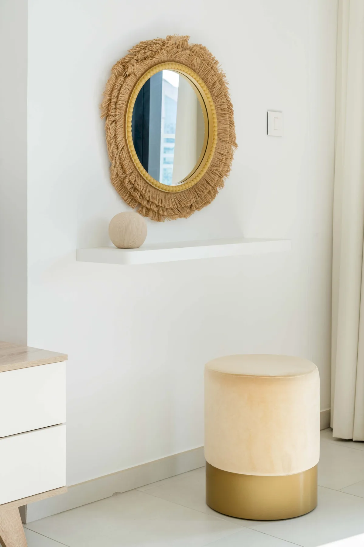 Una habitación luminosa con un taburete tapizado redondo bajo un espejo con flecos.