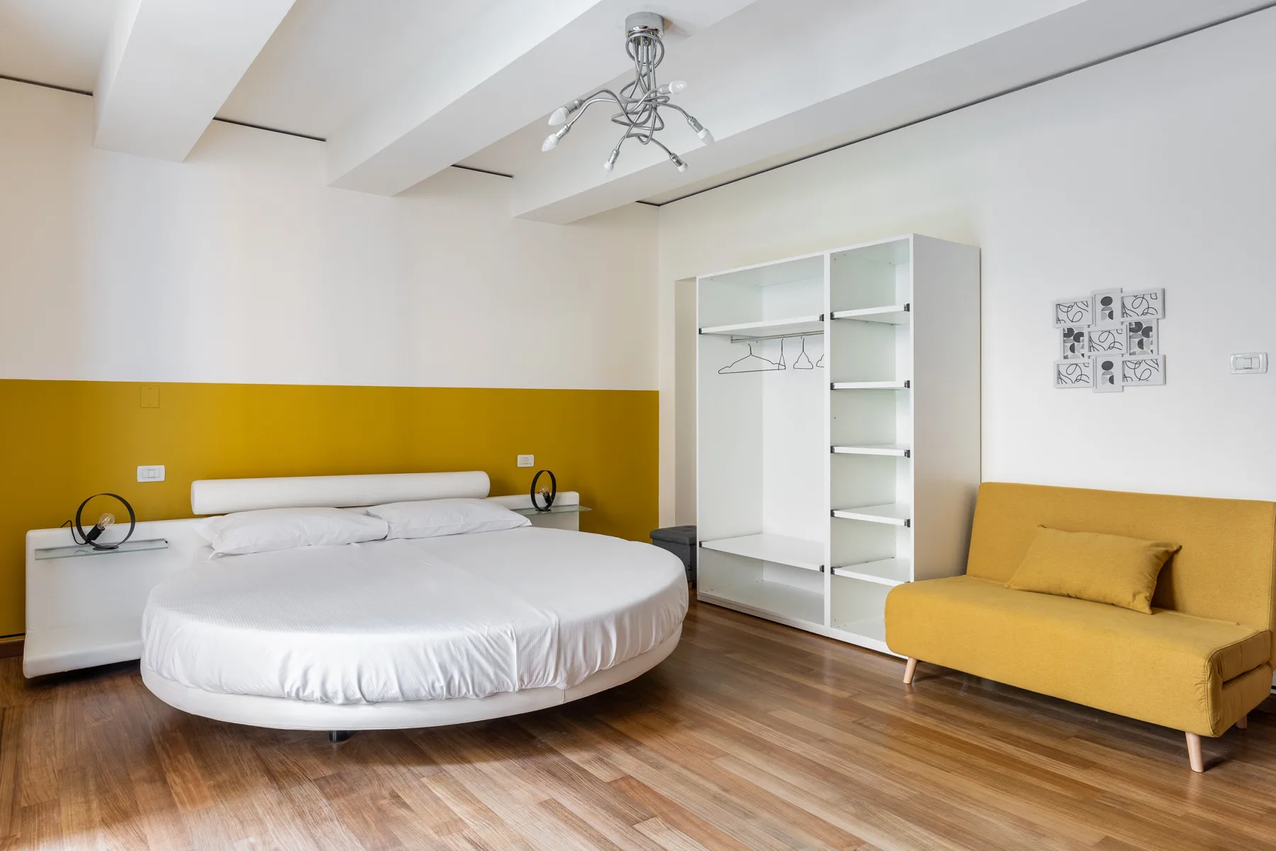A bedroom with a round bed, open wardrobe, and yellow seating.