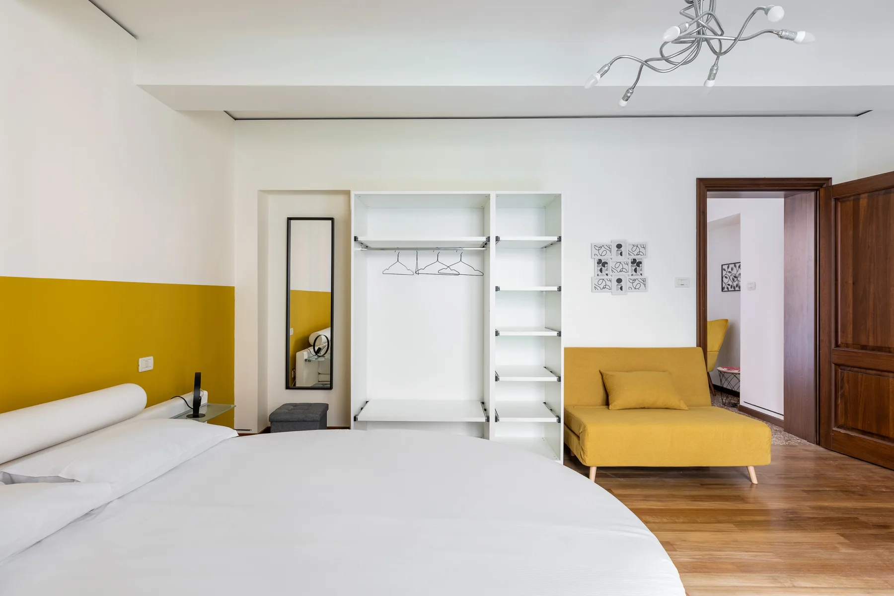 A bedroom with a bed, open wardrobe, and mustard-yellow seating.