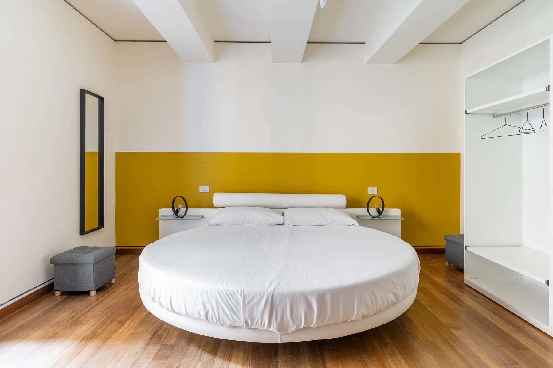 Un dormitorio luminoso con una cama redonda, mesitas de noche y una franja mostaza en la pared.