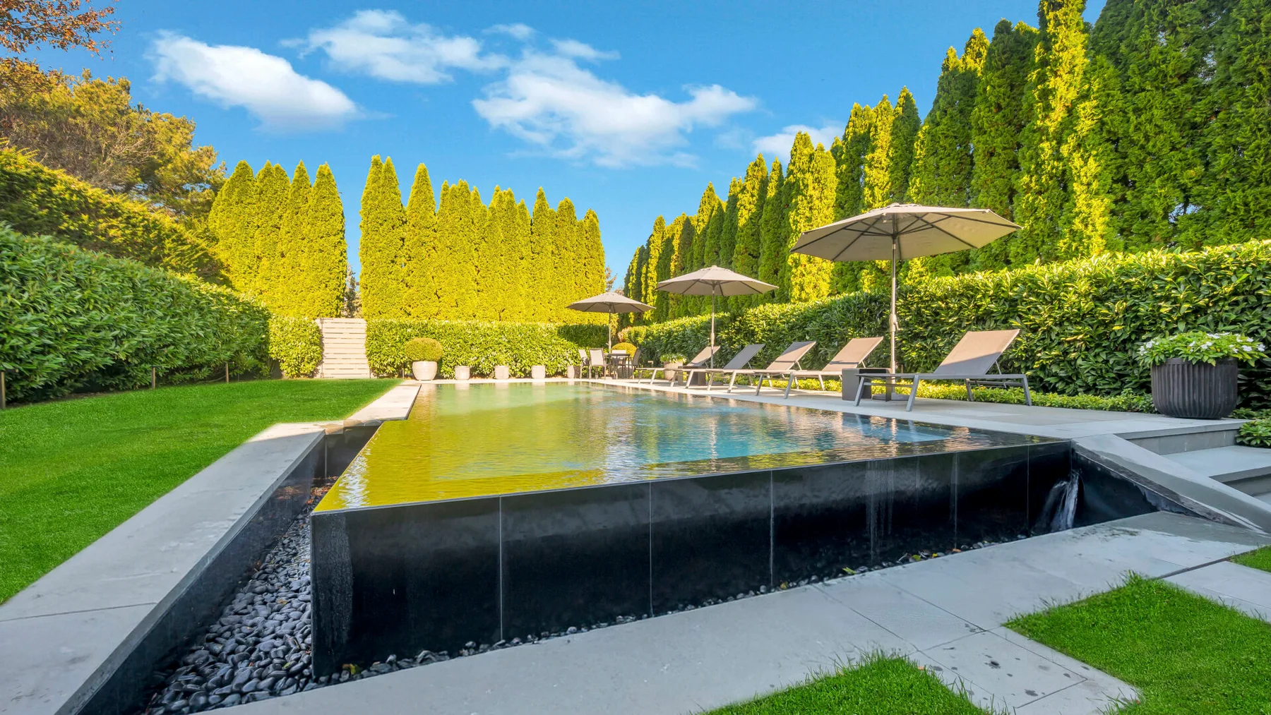 An outdoor swimming pool sits in a landscaped garden with lounge chairs and umbrellas.