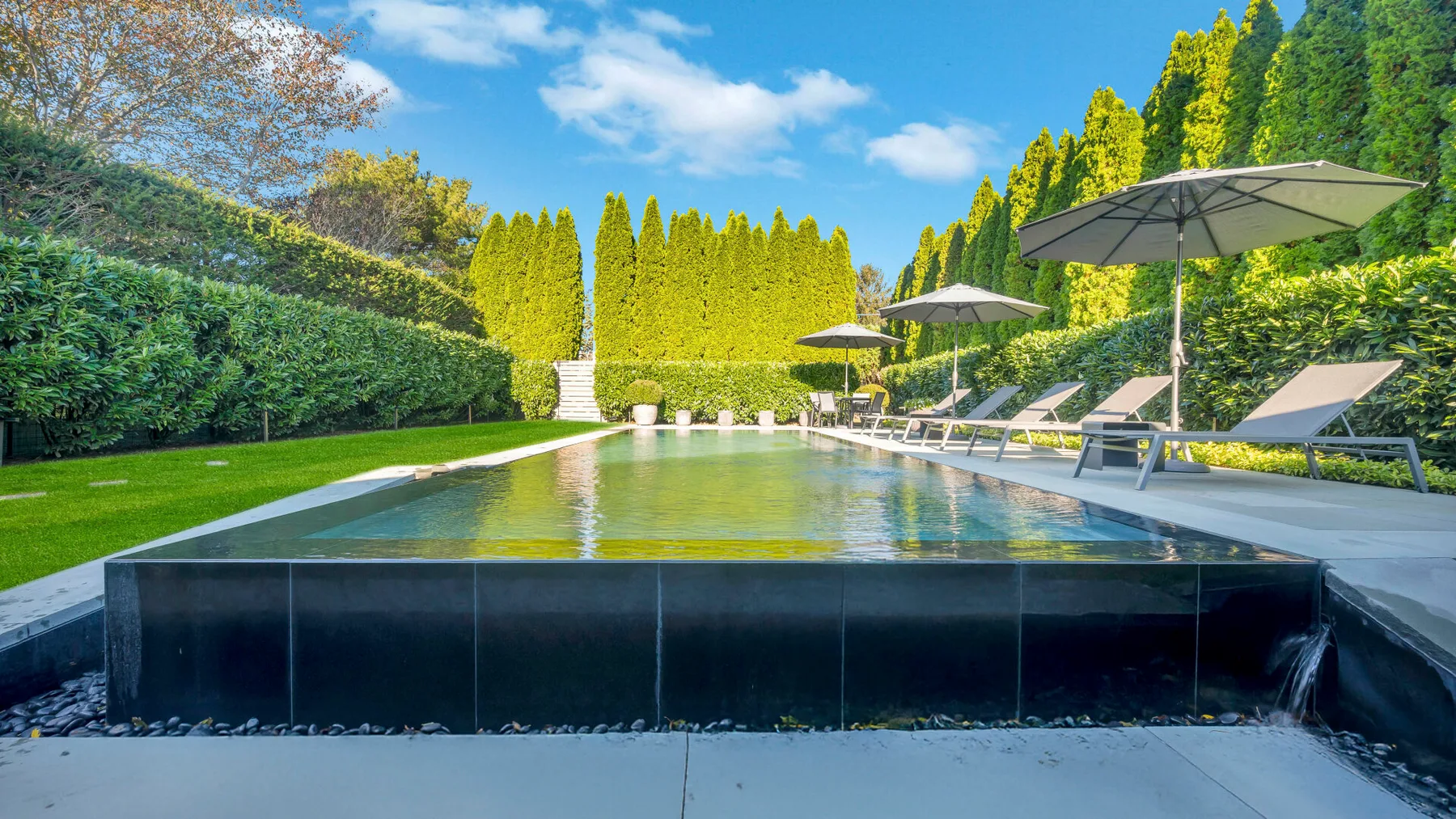 An outdoor swimming pool sits beside lounge seating and umbrellas in a green garden.